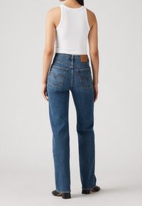 Kvinna som bär högmidjade raka blå jeans och en vit ärmlös topp, stående vänd bort på en enfärgad bakgrund.