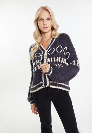 Granatowy sweter z dzianiny z białymi geometrycznymi wzorami, z oversize'owymi rękawami i dużymi guzikami. Podkreślony fakturowanym wzorem dzianiny.