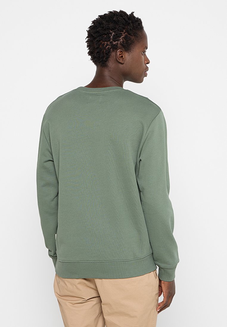 Personne avec des cheveux bouclés courts portant un sweat-shirt à manches longues vert et un pantalon beige, de dos, devant un fond blanc.
