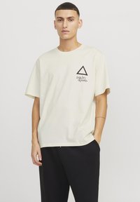 Camiseta de algodón beige con un logotipo triangular negro y el diseño del texto "SQUID GAME", combinada con pantalones negros. Mangas cortas y corte holgado.