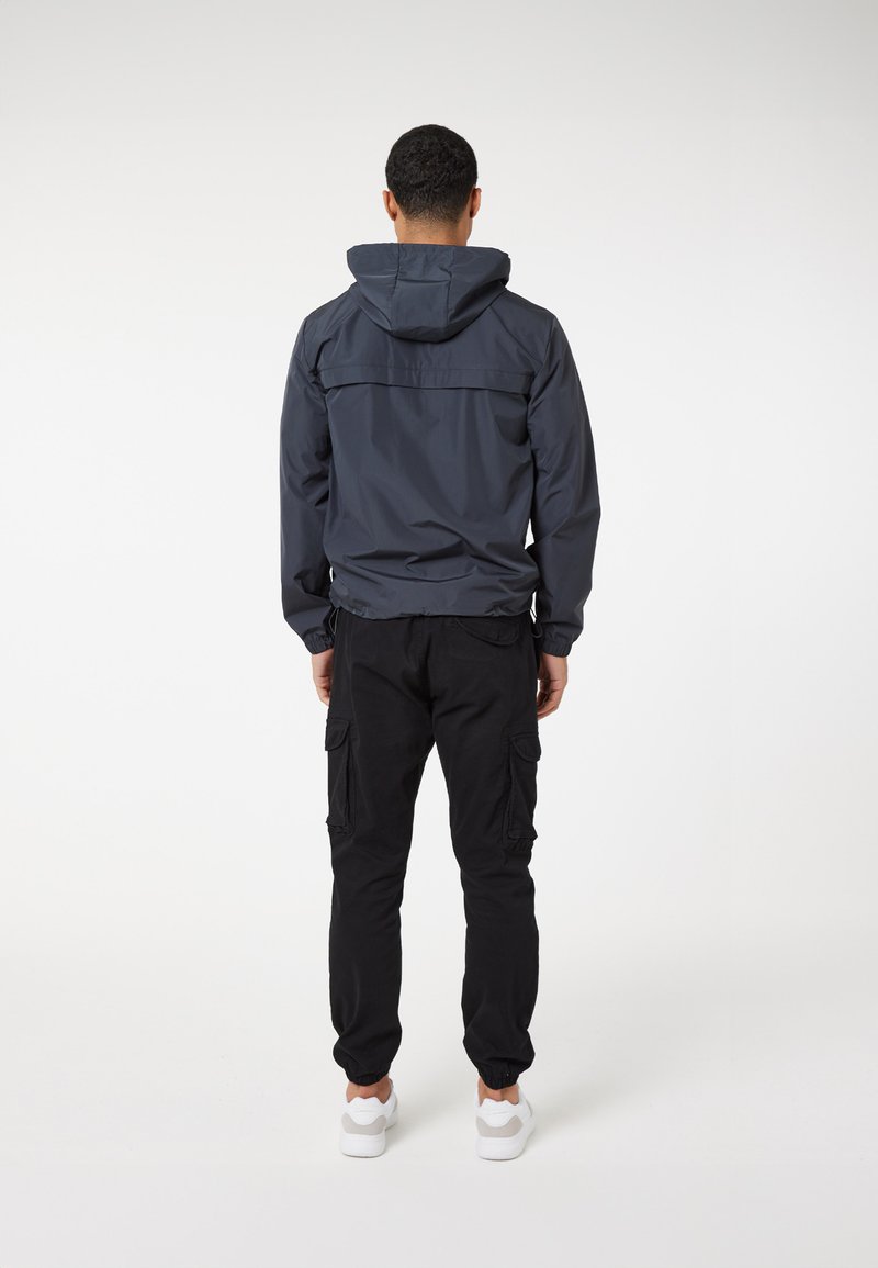 Blauwe hooded jacket met een gladde textuur en elastische manchetten, gecombineerd met zwarte cargobroek met zijzakken en elastische enkels. Witte schoenen.