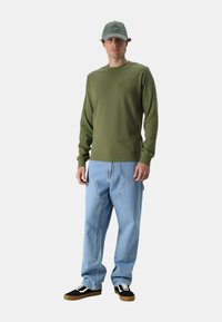 Camicia a maniche lunghe verde oliva in tessuto strutturato, jeans larghi di colore blu chiaro e scarpe da ginnastica nere con suole di gomma. Cappello di un morbido grigio.