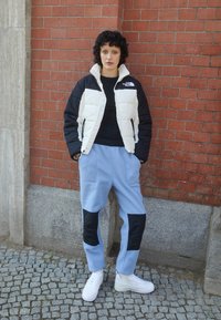 Padded jacket v čierno-bielej farbe, modré fleecové nohavice s čiernymi akcentmi a biele tenisky. Postáva proti textúrovanej tehlovej stene.