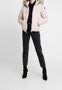 Světlounce růžová péřová bunda s kapucí lemovanou kožešinou, zapínáním na zip a elastickými manžetami. Kombinována s černými džínami s vysokým pasem a černými kotníkovými botami na podpatku.