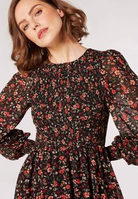 Blumenkleid mit schwarzem Hintergrund, das ein gesmocktes Oberteil, durchsichtige Puffärmel und ein Muster aus roten, rosa und grünen Blumen zeigt.