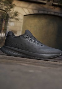 Black athletic running shoe with laces and thick cushioned sole, shown outdoors on stone ground with blurred urban background.