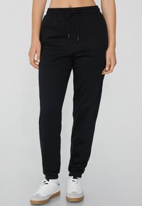Schwarze Jogginghose mit Kordelzug, schmal zulaufendem Bein und umgeschlagenen Säumen, aus einem weichen, leichten Material. Kombiniert mit weißen Sportschuhen.