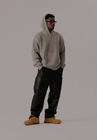 Modelo que lleva una sudadera de textura de gofre en gris claro, pantalones oversized negros y botas beige. La sudadera cuenta con un bolsillo frontal y un detalle de cremallera.