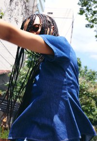 Personne aux longs cheveux tressés et lunettes de soleil, portant un haut bleu, tend le bras en plein air avec des arbres et des bâtiments en arrière-plan.