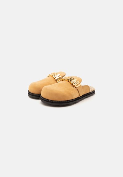Paire de sabots en daim beige à enfiler avec semelles noires et ornements de lettres dorées sur les brides supérieures sur fond blanc.