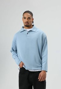 Hellblaues Langarm-Poloshirt mit Kragen, zwei schwarzen Knöpfen und lockerem Schnitt, getragen über einem weißen T-Shirt und kombiniert mit schwarzen Hosen.