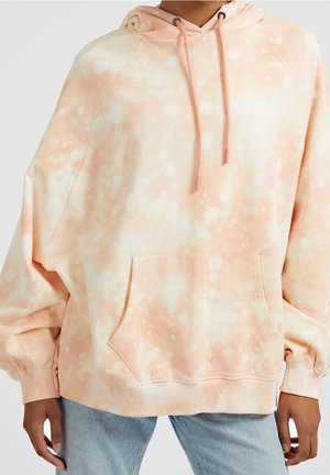 Osoba nosząca oversize bluzę z kapturem w brzoskwiniowo-białe wzory tie-dye z przednią kieszenią i dopasowanymi sznurkami, w połączeniu z jasnoniebieskimi jeansami.