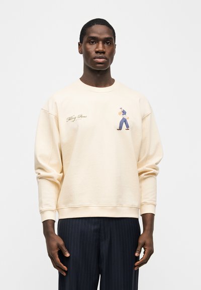 Άνδρας που φοράει μπεζ φούτερ με μικρό γραφικό περπατώντας πρόσωπο και κείμενο, συνδυασμένο με σκουρόχρωμο παντελόνι με λεπτές ρίγες, στέκεται μπροστά σε λευκό φόντο.