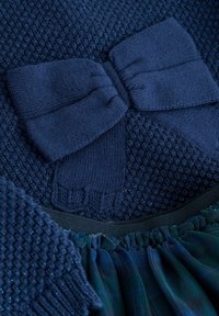 Navyfarbener Strickoberteil mit strukturiertem Muster und großem Schleifendetail. Der Saum ist fransig mit geschichtetem dunkelblauem Stoff und elastischem Bund.