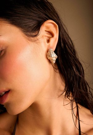 Ženska z mokrimi temnimi lasmi nosi teksturiran zlati uhan v obliki školjke na levem ušesu na nevtralnem ozadju.