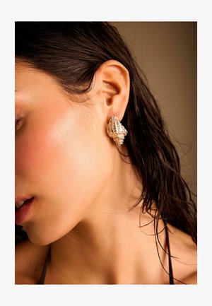 Donna con capelli scuri bagnati che indossa un orecchino di forma conchiglia dorata e testurizzata sull'orecchio sinistro, su sfondo neutro.