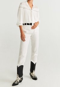 Weiße Bluse mit transparenten Einsätzen und Knöpfen, kombiniert mit weißen Hosen und schwarzen Cowboy-Stiefeln mit Schlangenlederdesign.