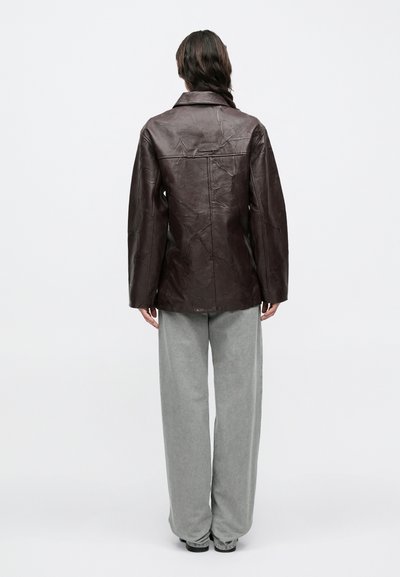 Bruine leren jas met een gekreukte textuur, kraagontwerp en rechte snit. Gecombineerd met loszittende grijze broek voor een ontspannen uitstraling.