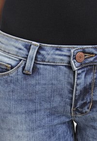 Vaaleansiniset denimfarkut, joissa on kevyt kulutus; mukana pronssinvärinen nappi, vyölenkit ja etutaskut. Rakenteellinen kangas, jossa näkyvät saumat.