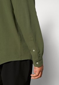 Oliwkowa koszula z długim rękawem o gładkiej fakturze, z zapięciem na guziki na mankietach oraz subtelnym ukośnym szwem w pobliżu dołu.