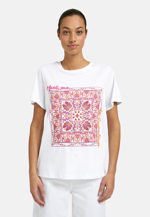 Woman wearing white t-shirt with pink and orange floral square pattern and text "Meet me" on upper left and right side.