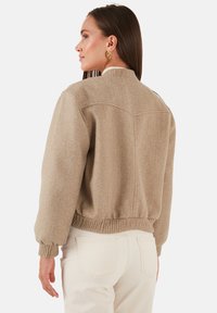 Beige gyapjú keverék bomber dzseki bordás mandzsettákkal és aljjal, kerek gallérral és hátsó yoke részlettel, krémszínű nadrággal párosítva.