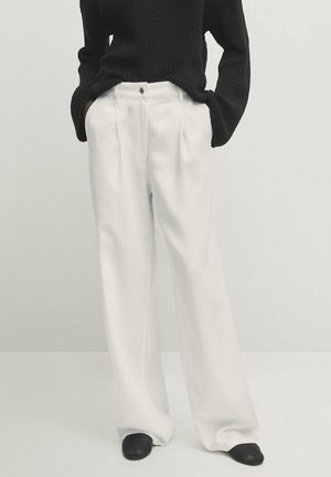 Personne portant un pantalon blanc ample plissé, un pull en maille noir, des chaussures noires, les mains dans les poches, sur un fond uni.