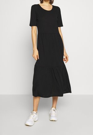 Woman wearing a black midi dress with short sleeves and a gathered hem, paired with white athletic sneakers, standing against a plain background.