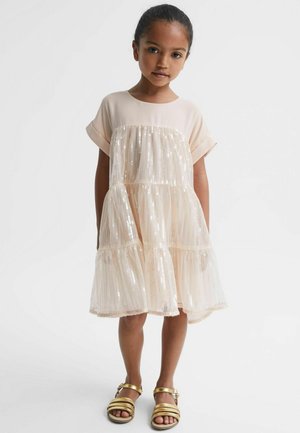 Noor tüdruk kannab helebeeži litrilist kihilist kleiti lühikeste varrukatega ja kuldseid sandaale, seisab ühevärvilise heleda tausta ees.