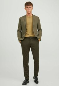 Traje ajustado verde oliva con un par de pantalones a juego, usado sobre un suéter marrón claro; presenta zapatos negros elegantes y un diseño minimalista.