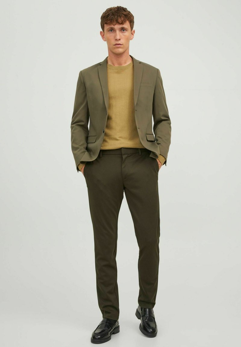 Traje ajustado verde oliva con un par de pantalones a juego, usado sobre un suéter marrón claro; presenta zapatos negros elegantes y un diseño minimalista.