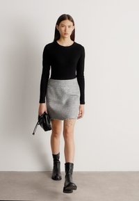 Νεαρή γυναίκα που περπατάει, φοράει μαύρο τοπ με μακριά μανίκια, γκρι μίνι φούστα από τουίντ, μαύρα μποτάκια μέχρι τον αστράγαλο, κρατώντας μια μαύρη τσάντα στο αριστερό της χέρι.