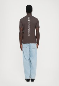 Vyras, vilkintis rudos spalvos Emporio Armani marškinėlius su vertikaliu baltu logotipu ant nugaros, šviesiai mėlynus džinsus ir juodus batus, stovi atsisukęs atgal.