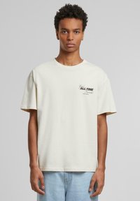 T-shirt beige à col rond en coton, avec des manches courtes et un détail de texte imprimé. Porté avec un jean en denim bleu clair.