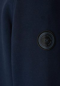 Dunkelblaues strukturiertes Gewebe mit gerippter Musterung, versehen mit einem runden schwarzen Logo-Patch mit blauen Akzenten auf der linken Schulter.