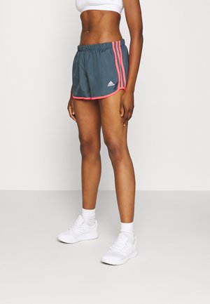 Modelo femenina con pantalones cortos deportivos grises de Adidas con rayas y ribetes rosas, sujetador deportivo blanco, calcetines blancos y zapatillas blancas, de pie frente a un fondo blanco.