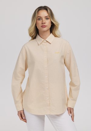 Kobieta o blond włosach, ubrana w beżowo-białą, paskowaną koszulę na guziki z długim rękawem oraz białe spodnie, patrzy przed siebie z neutralnym wyrazem twarzy.