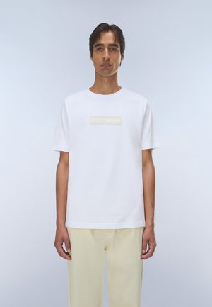 Ung mand med kort mørkt hår iført en hvid kortærmet skjorte med et svagt beige logo og lyse beige bukser mod en enkel baggrund.
