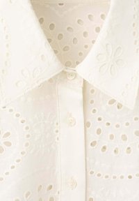 Blouse blanche boutonnée avec broderie florale en œillet et col pointu.