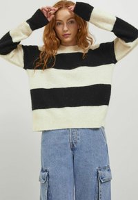 Maglione a righe nere e crema con una consistenza morbida, dotato di maniche lunghe e collo a giro ampio, abbinato a jeans cargo blu chiaro.
