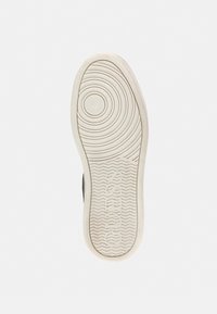 Semelle de chaussure en caoutchouc blanc avec des motifs circulaires et ondulés, portant le nom de la marque "GUESS" embossé près du talon. Texture lisse dans l'ensemble.