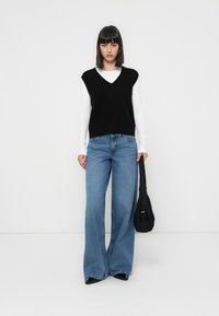 Zwarte mouwloze trui gedragen over een witte blouse met lange mouwen, gecombineerd met lichtblauwe flared jeans en een zwarte tas.