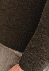 Ciemnozielona dzianina swetra z prążkowanymi mankietami i dołem, charakteryzująca się miękką teksturą i gładkim wykończeniem. Zbliżenie na szczegóły.