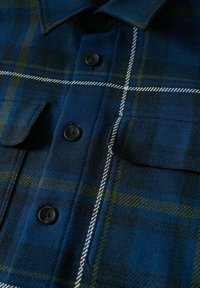 Camicia a quadri blu e verde con colletto a punta, due tasche sul petto, bottoni neri e dettagli in cucitura bianca su un morbido tessuto testurizzato.