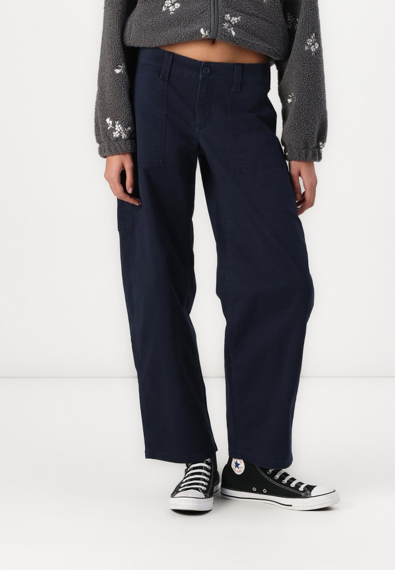 Calças largas azul marinho com fecho de botão, apresentando bolsos laterais e uma textura de algodão suave, combinadas com ténis pretos.