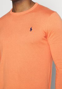 Ljust orange tröja, tillverkad av mjukt material, har en rund hals och en liten marinblå logotyp av en häst på vänster bröst.