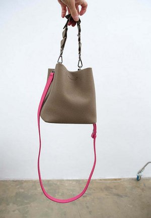 Kbelíková taška z hnědé texturované kůže s kontrastním růžovým popruhem. Má krátkou rukojeť a dlouhý odnímatelný popruh, minimalistický design.
