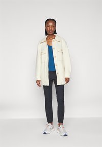 Crème wollen blend jas met grote zakken, knoopsluiting; gecombineerd met een blauwe top, zwarte tapered broek en witte sportschoenen.