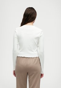 Biała bluzka z długim rękawem o prążkowanej teksturze i falbaną na dole, w połączeniu z jasnobrązowymi spodniami w luźnym fasonie. Widok z tyłu przedstawia minimalne detale.