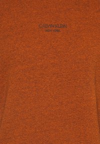 Tessuto a maglia arancione con texture che presenta il logo "Calvin Klein New York" in un colore scuro a contrasto. Design semplice, peso medio.